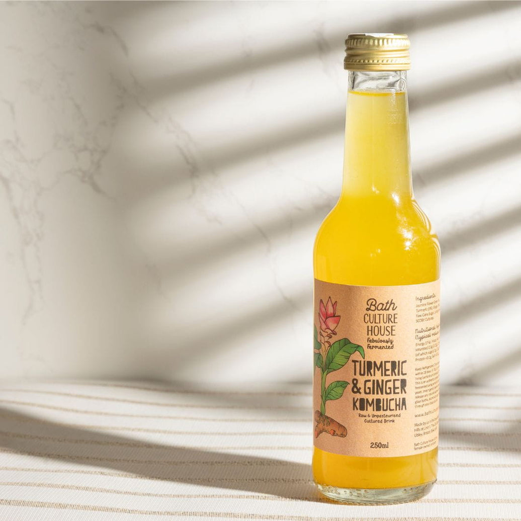 Bottle of Bath Culture House Turmeric & Ginger Kombucha on a marble surface with sunlight filtering through.