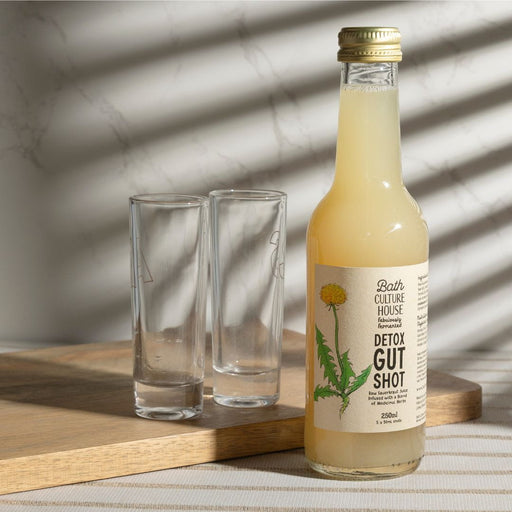 Bottle of Bath Culture House Detox Gut Shot with two glasses on a wooden surface.