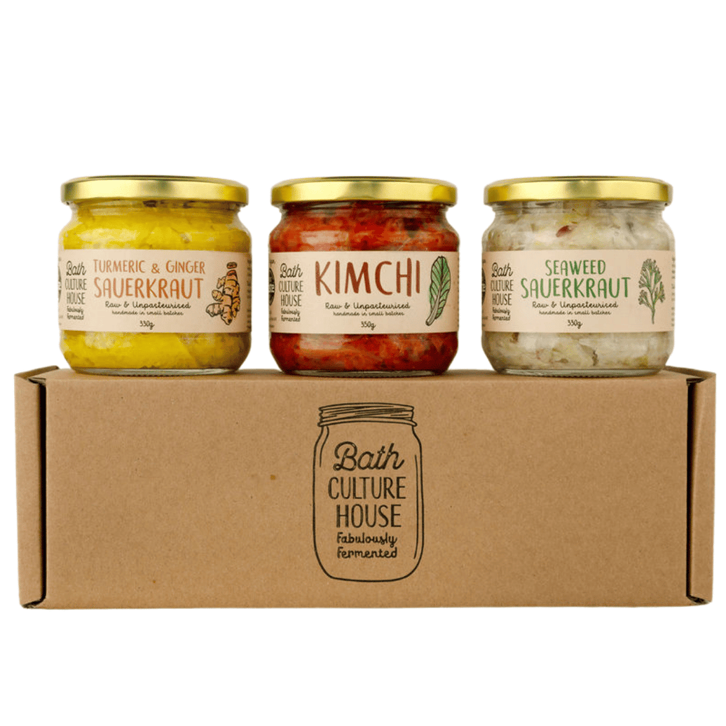 Three glass jars of Bath Culture House fermented foods with gold lids, displayed on top of a brown cardboard gift box. The jars contain turmeric and ginger sauerkraut, kimchi, and seaweed sauerkraut.