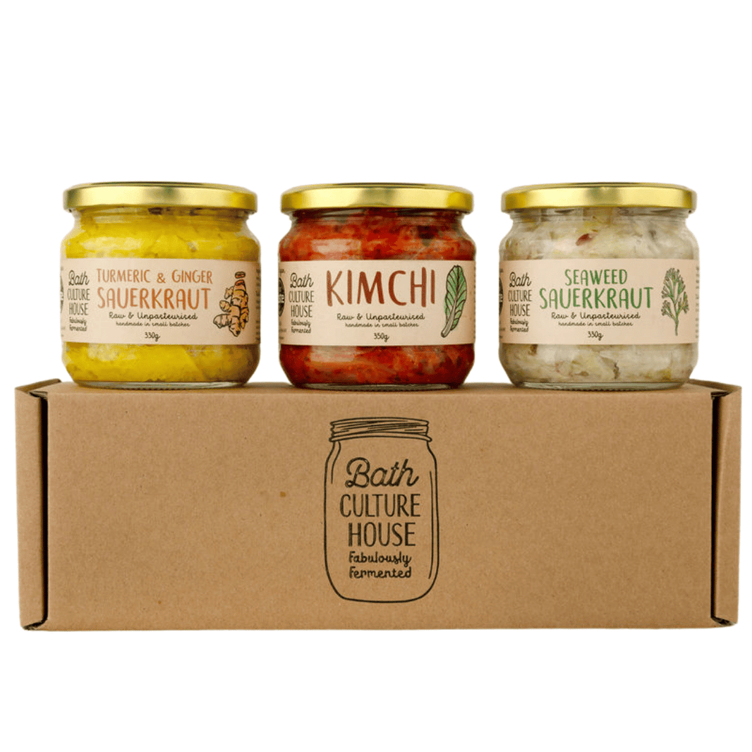 Three glass jars of Bath Culture House fermented foods with gold lids, displayed on top of a brown cardboard gift box. The jars contain turmeric and ginger sauerkraut, kimchi, and seaweed sauerkraut.