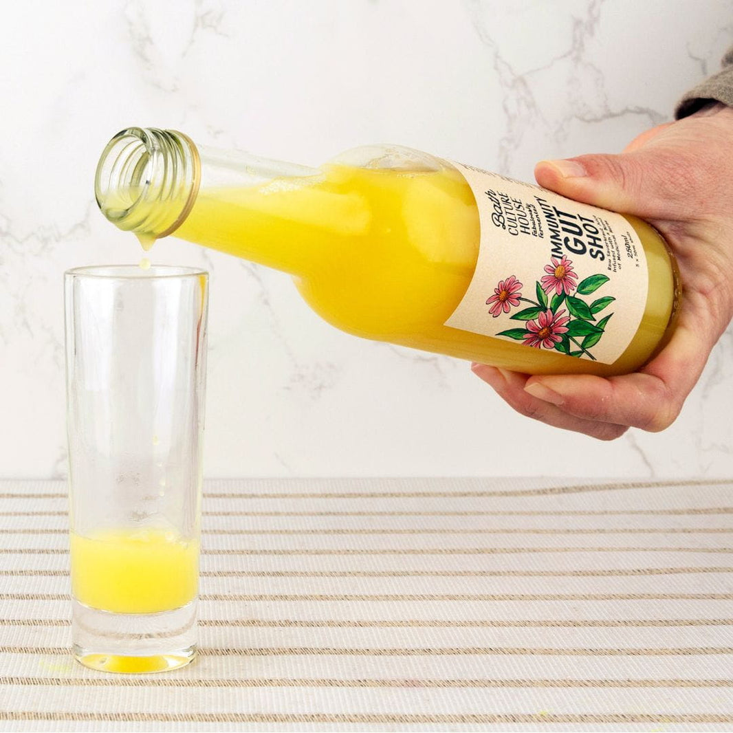 Hand pouring a yellow liquid from a bottle labeled 'Gut Shot' into a glass on a marble surface.