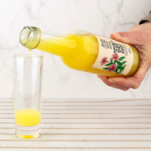 Hand pouring a yellow liquid from a bottle labeled 'Gut Shot' into a glass on a marble surface.