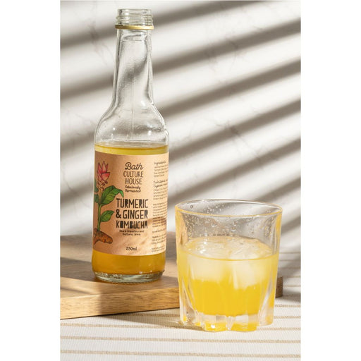 Bottle of Bath Culture House Turmeric & Ginger Kimchi with a glass of the drink on a striped surface.